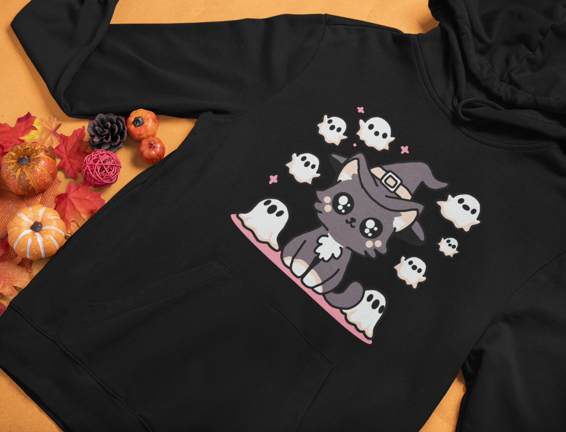 Black hoodie with a Halloween-themed design of a cat in a witch hat surrounded by ghosts on a wooden surface.