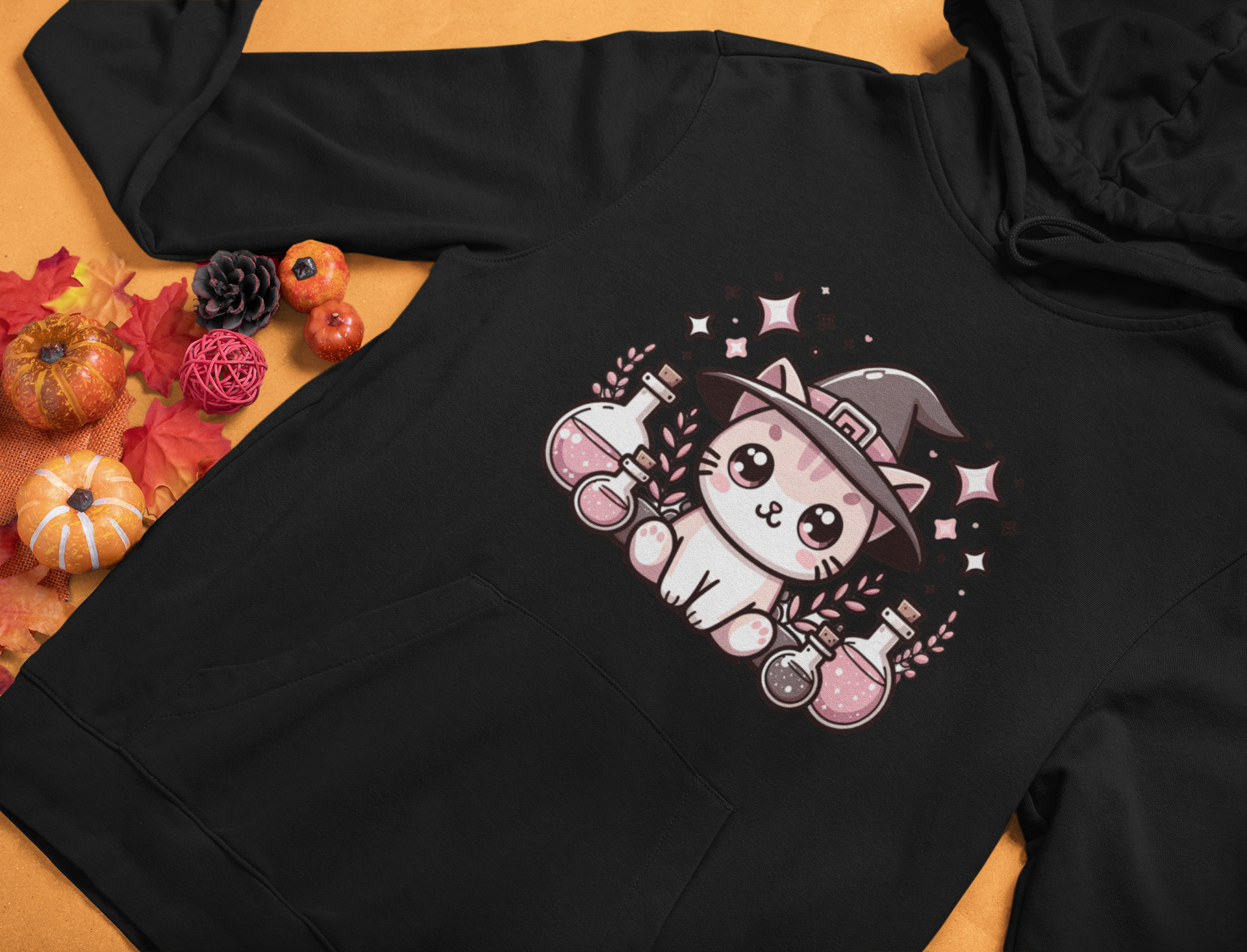 Black hoodie with a cute cat in a witch hat design on a wooden surface with fall decorations.